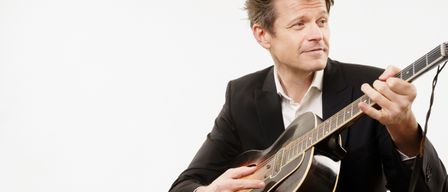 Ein Mann in einem Anzug spielt eine Gitarre und schaut in die Kamera. Die Gitarre hat einen goldbraunen Korpus und einen schwarzen Hals.