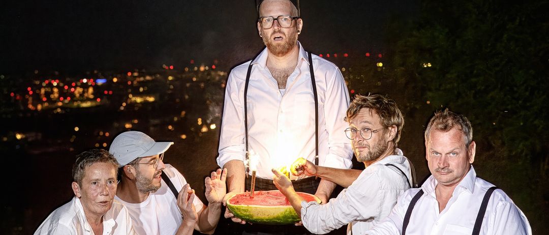 Fünf Männer in weißen Hemden und Hosenträger posieren mit einer Wassermelone. Sie halten Wunderkerzen und stehen vor einer Stadtsilhouette.