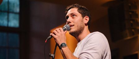 Ein Mann mit Bart singt in ein Mikrofon auf der Bühne. Er trägt ein graues Hemd und eine Uhr am linken Handgelenk.