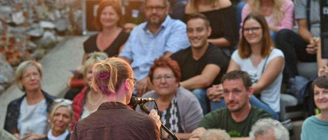 Eine Frau steht vor einem Mikrofon und spricht zu einer Menschenmenge, die aus verschiedenen Personen besteht, einige sitzend, einige stehend, alle konzentrieren sich auf sie. Ein helles Licht beleuchtet sie von links.