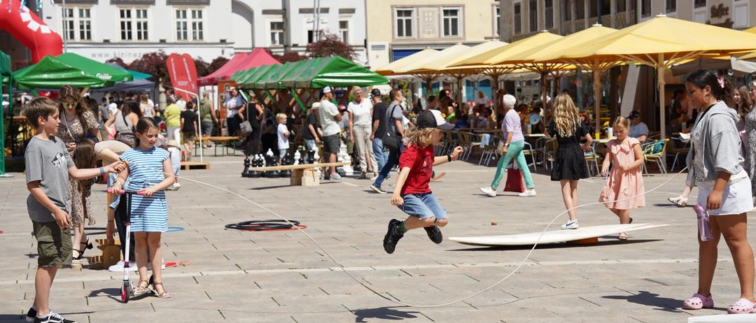 Ein Junge springt über ein Seil, das ein Mädchen hält. Sie befinden sich in einem geschäftigen Außenbereich mit vielen Menschen, Geschäften und Sonnenschirmen. Gebäude mit vielen Fenstern umgeben den Bereich.