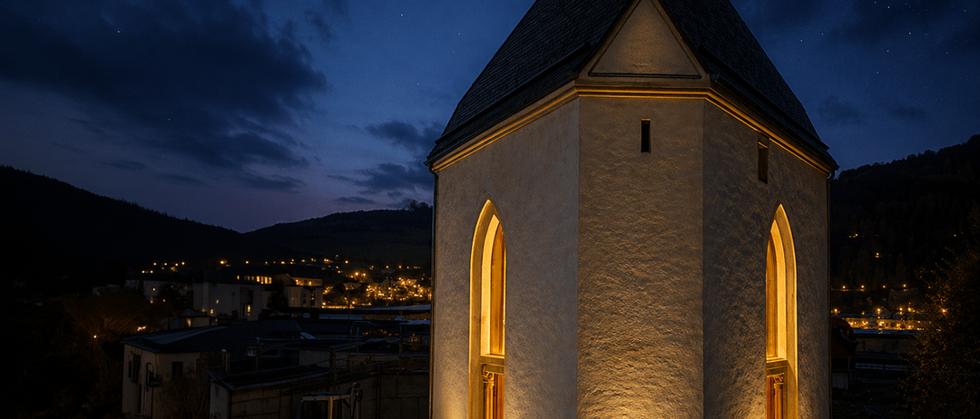 Eine Kapelle mit einem dreieckigen Dach steht beleuchtet in der Nacht. Das Gebäude wird von warmen gelben Lichtern beleuchtet, wobei die Stadtlichter in der Ferne schwach sichtbar sind.