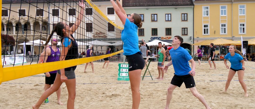 Eine Gruppe von Menschen spielt Volleyball auf einem Sandplatz mit einem gelben Netz. Ein Spieler springt, um den Ball zu schlagen. Im Hintergrund befinden sich Gebäude mit Schildern.