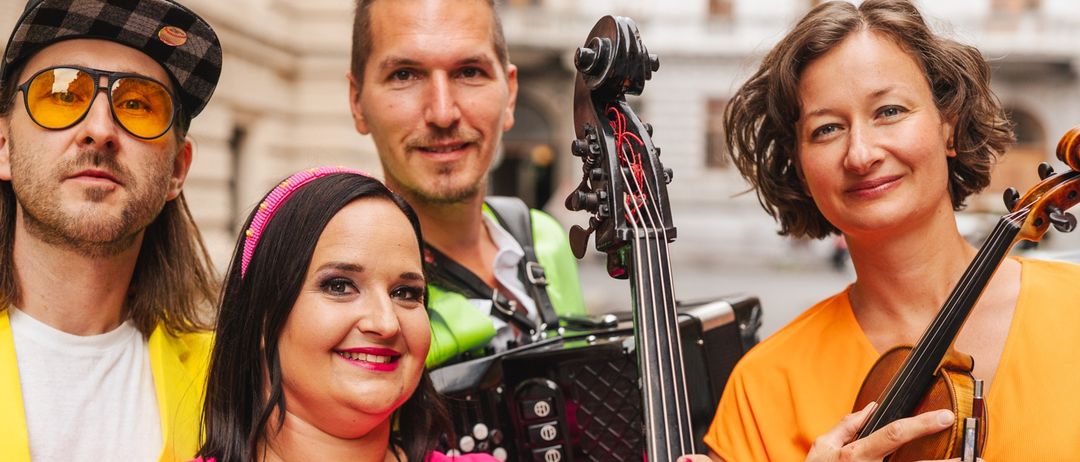 Eine Gruppe von Musikern posiert für ein Foto. Die Frau in Pink hält eine Geige, während die Frau in Orange eine Geige hält. Ein Mann spielt Akkordeon.