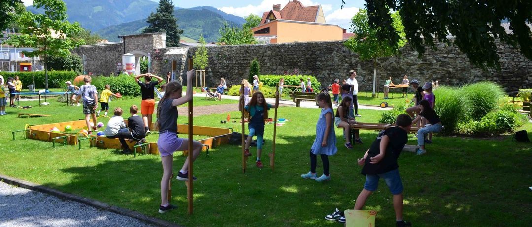 Kinder spielen auf einem Rasenfeld mit einer Steinmauer im Hintergrund, Berge in der Ferne.