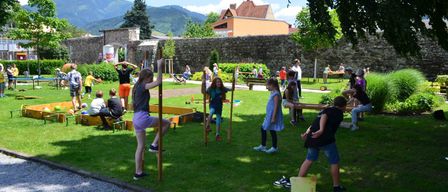 Kinder spielen auf einem Rasenfeld mit einer Steinmauer im Hintergrund, Berge in der Ferne.