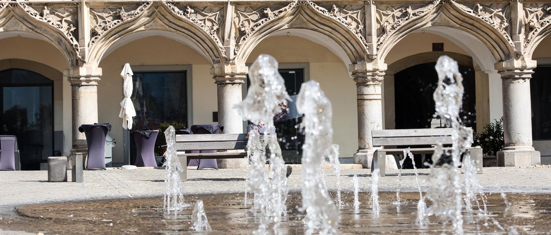 Ein Brunnen vor einem historischen Gebäude mit verzierten Bögen und Balkonen, mit kunstvollen Steinarbeiten und moderner Architektur.