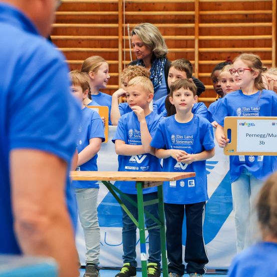 Kinder in blauen Shirts stehen um einen Tisch, einer hält ein Schild. Eine Frau steht hinter ihnen. Sie sind möglicherweise in einem Wettbewerb.