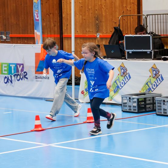 Zwei Kinder in blauen T-Shirts spielen eine Runde Fangen in einer Turnhalle, mit orangefarbenen und weißen Hütchen als Spielgrenzen.