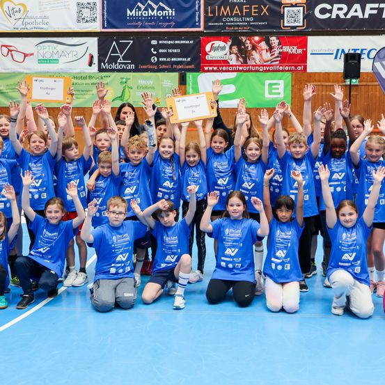 Eine Gruppe junger Kinder in blauen T-Shirts mit einem Zertifikat posiert für ein Foto in einer Turnhalle. Dahinter befinden sich Plakate und Poster.