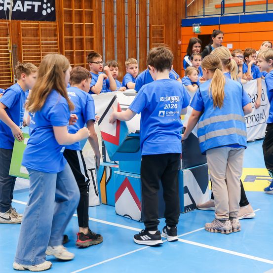 Eine Gruppe junger Kinder in blauen T-Shirts versammelt sich um einen bunten Hindernisparcours in einer Turnhalle. Einige tragen Sicherheitswesten. Dahinter beobachten andere Kinder.