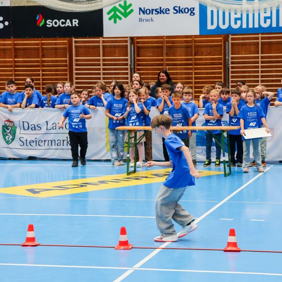 Eine Gruppe von Kindern in blauen Shirts nimmt an einem Sportereignis auf einem Platz mit orangefarbenen Kegeln teil. Ein Erwachsener hält ein Papier vor ihnen.