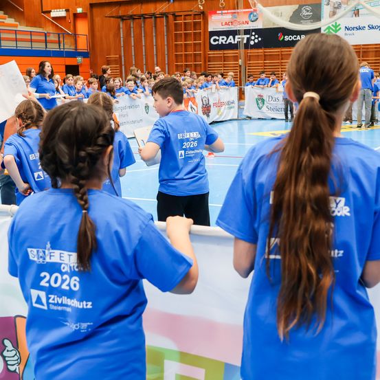 Eine Gruppe von Kindern in blauen Uniformen mit Nummern steht in einer Sporthalle. Ein Junge spricht mit einem Mädchen mit einem Pferdeschwanz.