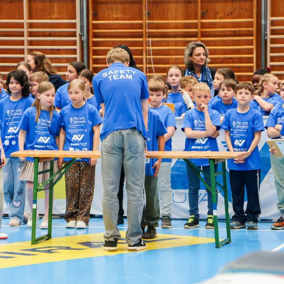 Eine Gruppe von Kindern in blauen T-Shirts mit der Aufschrift 'Sicherheitsteam' steht hinter einem Holztisch. Ein Erwachsener steht vor ihnen und bereitet möglicherweise eine Präsentation vor.