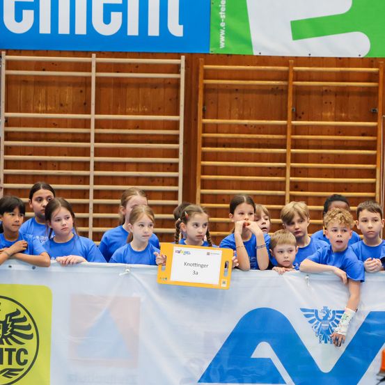 Kinder in blauen Shirts stehen hinter einem Banner mit 'ATC' und einem Logo. Eines hält ein gelbes Namensschild, 'Knottinger 3a.' Sie blicken auf etwas jenseits der Barriere.