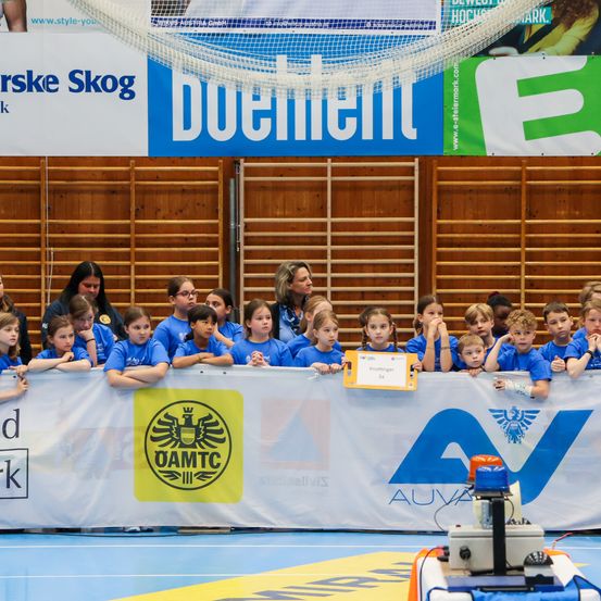 Kinder in blauen Shirts beobachten eine Veranstaltung und stehen hinter einem Banner mit verschiedenen Logos und einem gelben Schild. Die Wand dahinter hat Holzstrukturen und Banner.