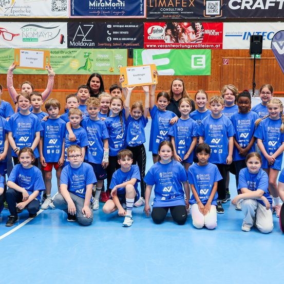 Eine Gruppe von Kindern in blauen T-Shirts, wahrscheinlich von einem Sportteam, posiert für ein Foto auf einem blauen Boden. Eine Frau vor ihnen hält ein gelbes Papier. Hinter ihnen sind Banner mit Logos und Texten.