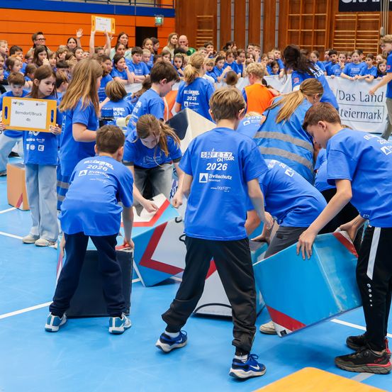 Eine Gruppe von Kindern in blauen Uniformen nimmt in einer Turnhalle an einer Team-Building-Aktivität teil. Einige halten blaue Tafeln, während andere mit einer dreieckigen Struktur interagieren. Ein Publikum beobachtet im Hintergrund.