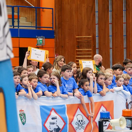 Eine Gruppe von Kindern in blauen T-Shirts schaut sich eine Veranstaltung an, bei der andere Schilder hochhalten, und einer Banner mit einem Symbol und Logo.