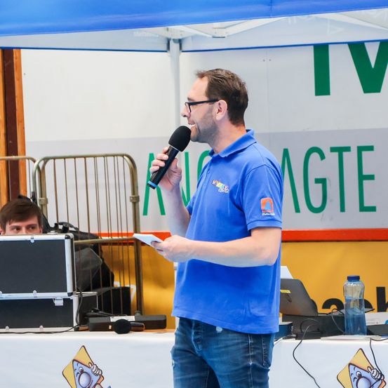 Ein Mann in einem blauen Polohemd steht unter einem Zelt an einem Mikrofon, hält ein Blatt Papier in der Hand, mit einer Wasserflasche und einem Laptop auf dem Tisch hinter ihm.