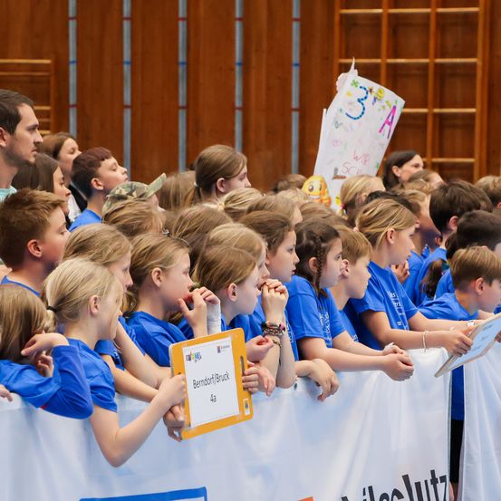 Eine Gruppe von Kindern in blauen Shirts steht zusammen. Sie halten ein Banner mit dem Wort 'Berndorf' darauf. Die Kinder schauen auf etwas vor ihnen.