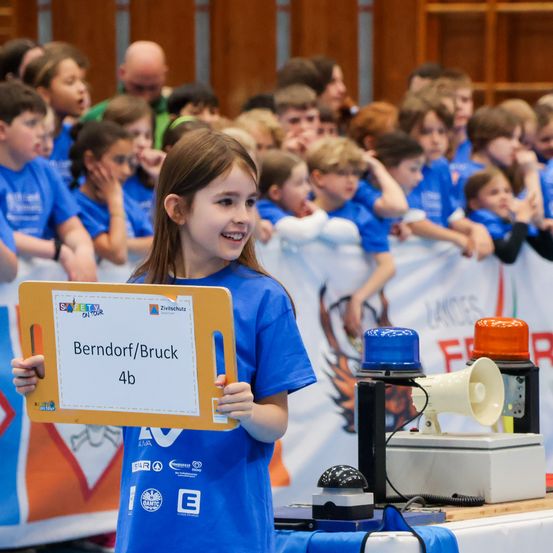 Ein Mädchen hält ein Schild mit Berndorf/Bruch 4b, vor einer Menschenmenge in blauen T-Shirts. Dahinter ist ein weißes Banner mit Logos zu sehen, und ein Tisch hält ein Megaphon und Sirenen.