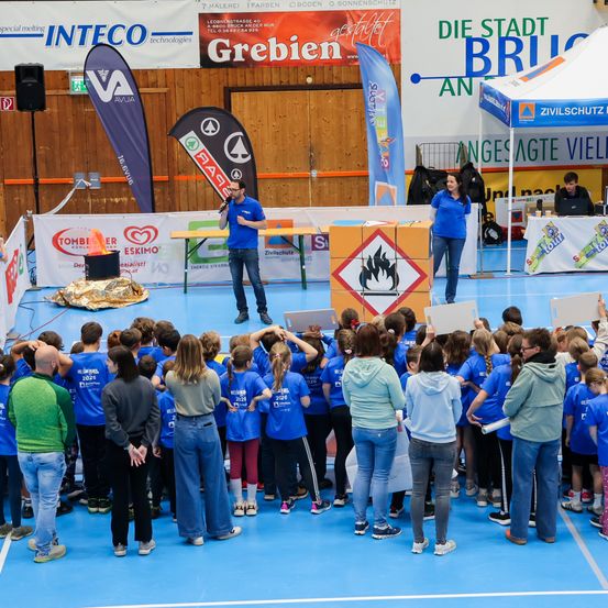 Eine Gruppe von Schülern in blauen Hemden hört einem Mann vor einer Bühne zu. Auf der Bühne stehen zwei Frauen und ein Mann. Vor einer Wand mit Text steht ein Zelt mit einem Tisch.