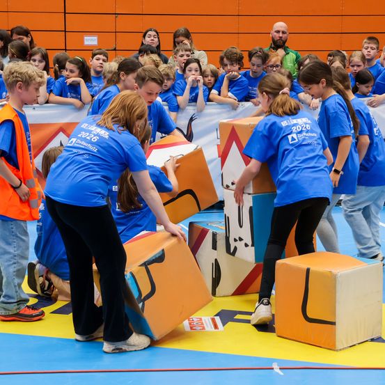 Kinder in blauen T-Shirts arbeiten an einem Projekt mit bunten Blöcken, beaufsichtigt von Erwachsenen, während Zuschauer hinter einer Barriere zusehen.