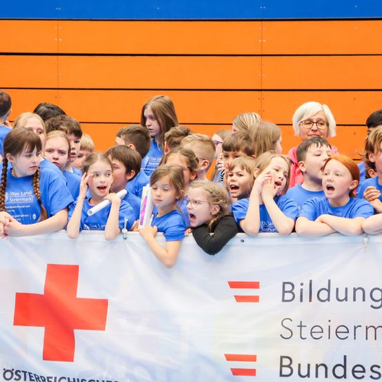 Eine Gruppe von Kindern in blauen T-Shirts steht hinter einem Banner, einige schreien, und sie scheinen an einer Schulveranstaltung teilzunehmen.