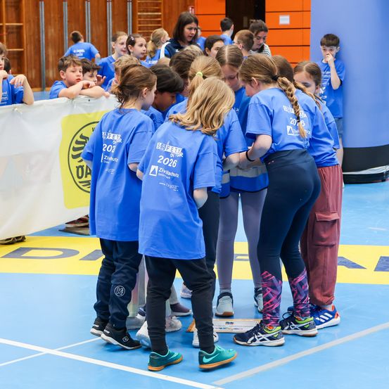 Gruppe von Kindern in blauen T-Shirts, die sich in einer Turnhalle versammelt und zuhören. Ein Banner und ein blauer Boden deuten auf eine Schulveranstaltung hin.
