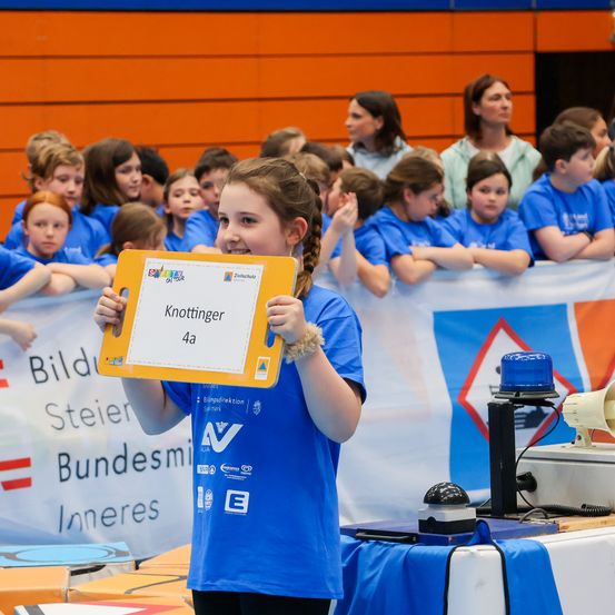 Ein Mädchen hält eine gelbe Plakette hoch, auf der Knottinger 4a steht, und steht vor einer Gruppe von Kindern in blauen Shirts. Der Hintergrund zeigt Banner mit Text auf Deutsch.