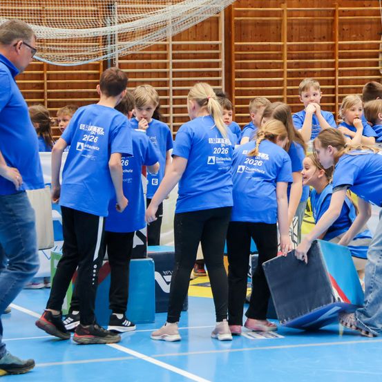 Gruppe von Kindern in blauen T-Shirts mit Nummern auf dem Rücken, die in einer Turnhalle trainieren. Einige halten blaue Objekte, während andere zuschauen.