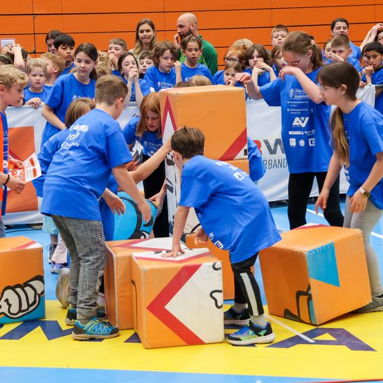 Eine Gruppe von Kindern in blauen T-Shirts spielt mit großen Würfeln auf einem farbenfrohen Boden. Einige tragen Brillen und Turnschuhe, während andere hinter einer Barriere zuschauen. Die Umgebung ist drinnen, mit Bannern und einer orangefarbenen Wand.