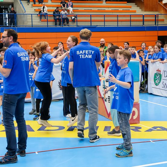 Mehrere Personen in blauen Sicherheitsteam-Shirts stehen in einer Sporthalle. Sie sprechen mit einem Jungen. Dahinter stehen Leute auf den Tribünen.