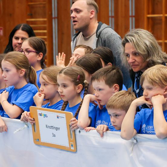 Eine Gruppe von Kindern in blauen T-Shirts schaut eine Veranstaltung, einige mit Brille. Ein Schild sagt Knottinger 3a. Eine Frau und ein Mann schauen auch zu.