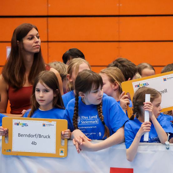 Eine Gruppe junger Mädchen in blauen T-Shirts hält ein gelbes Schild mit den Worten Berndorf/Bruck 4b. Sie stehen vor einer Barriere und schauen nach vorne. Hinter ihnen steht eine Frau mit langen Haaren. Im Hintergrund ist ein verschwommener Blick auf Menschen zu sehen, die vor einer Wand stehen.