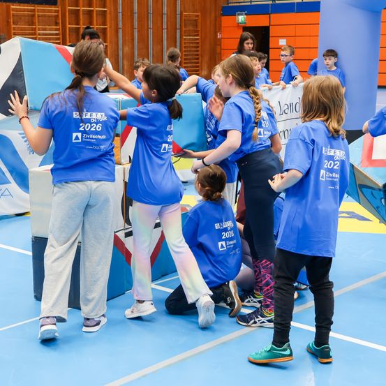 Eine Gruppe von Kindern in blauen T-Shirts mit der Nummer 2026 darauf spielen und halten sich gegenseitig auf einem blauen Boden fest. Dahinter sind andere Kinder und Erwachsene.