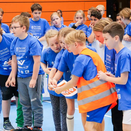 Eine Gruppe von Kindern in blauen T-Shirts und einigen mit Warnwesten versammelt sich auf einem Platz, vermutlich in Vorbereitung auf eine Veranstaltung. Ein Junge in der Mitte hält einen Ball und sieht ihn an.