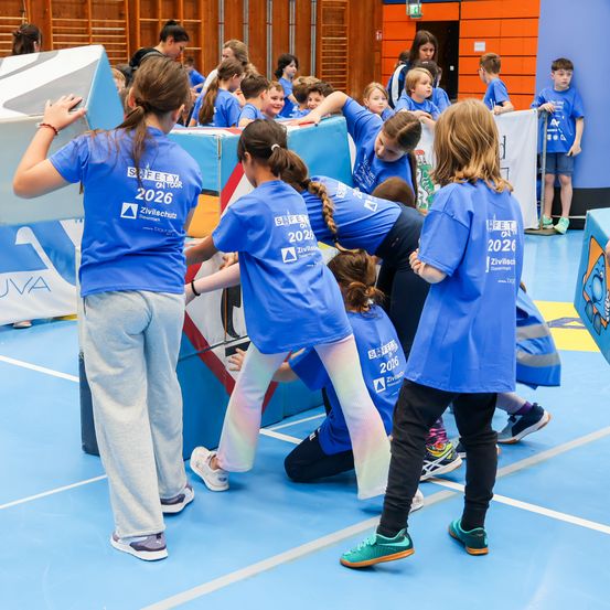 Eine Gruppe von Kindern in blauen T-Shirts mit 2026 darauf, die sich an einer Teambuilding-Aktivität in einer Turnhalle beteiligen. Sie stehen, beugen sich und schauen sich etwas an.