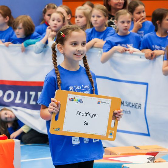 Ein Mädchen in einem blauen Shirt hält ein gelbes Schild mit dem Text Knotfinger 3a vor einer Gruppe von Kindern. Hinter ihr ist ein Banner mit dem Wort Polizei zu sehen.