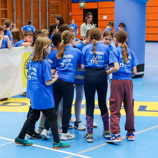 Eine Gruppe von Kindern in blauen T-Shirts mit der Nummer 2026 steht in einem Kreis in einer Turnhalle. Sie nehmen wahrscheinlich an einer Veranstaltung teil.