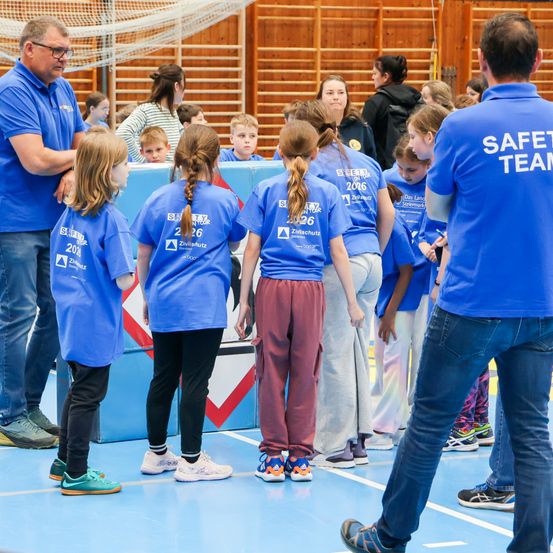Eine Gruppe von Kindern in blauen Hemden mit Nummern steht in einer Turnhalle, überwacht von Erwachsenen. Die Erwachsenen tragen blaue Hemden mit weißem Text. Eine Holzwand mit Kletterstangen ist im Hintergrund.