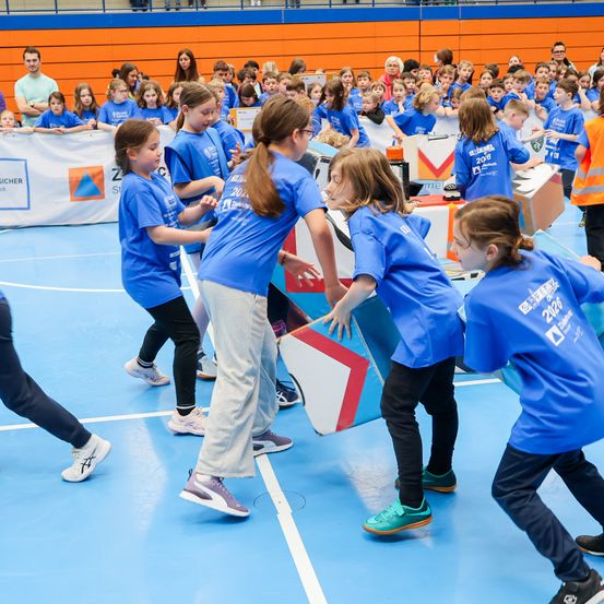 Junge Mädchen in blauen Uniformen spielen auf einem Sportplatz ein Spiel, während ein Publikum zusieht.