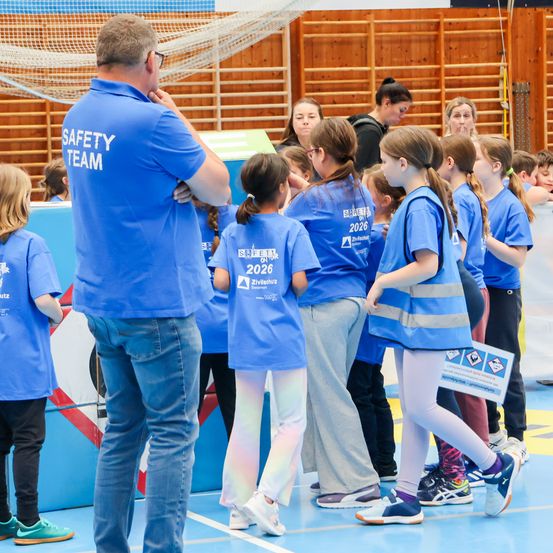 Eine Gruppe von Kindern in blauen Shirts steht in einer Turnhalle und hört einem Sicherheitsteammitglied zu. Die Kinder haben Nummern auf ihren Shirts, und das Sicherheitsteammitglied hat ein blaues Shirt mit der Aufschrift Sicherheitsteam.