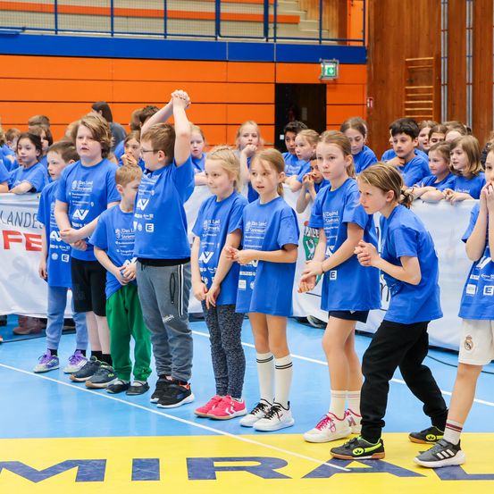 Kinder in blauen T-Shirts stehen auf einem blauen Boden, wahrscheinlich in einer Schulsporthalle, und heben die Hände. Einige tragen Turnschuhe, und ein Banner mit weißem Text hängt hinter ihnen.