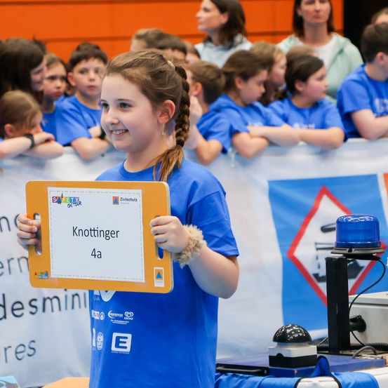 Ein Mädchen in einem blauen T-Shirt hält ein Schild hoch, auf dem Knottinger 4a steht, vor einem Publikum.