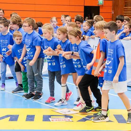 Gruppe von Kindern in blauen T-Shirts steht auf einem Sportplatz mit gelben Markierungen, einige tragen Brillen, Turnschuhe und Socken. Sie scheinen an einem Sportereignis teilzunehmen.