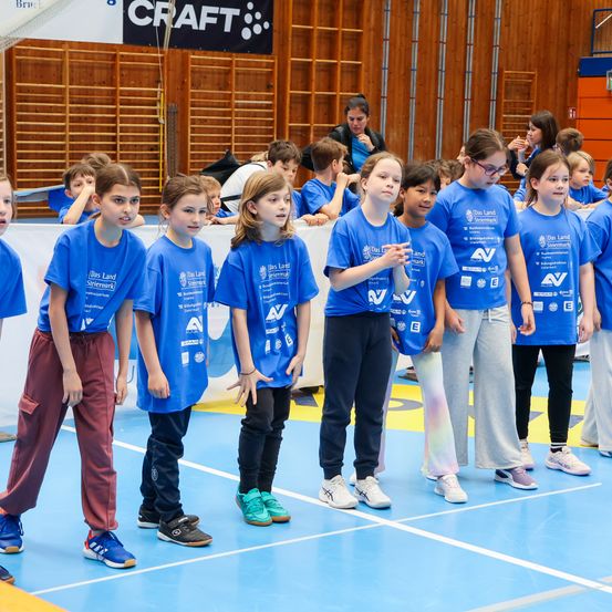 Eine Gruppe von Kindern in blauen T-Shirts mit einem Logo steht auf einem Sportplatz, vor einer Holzwand mit einem Banner. Sie warten scheinbar auf etwas.