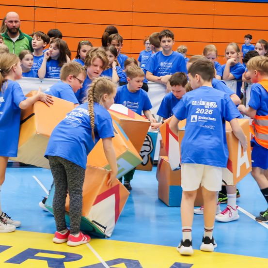 Eine Gruppe junger Schüler, die blaue T-Shirts mit dem Jahr 2026 bedruckt tragen, spielt ein Spiel auf einem blauen Boden.