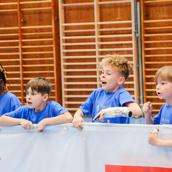 Vier junge Jungen in blauen T-Shirts lehnen sich über eine weiße Barriere. Sie alle schauen nach oben, möglicherweise bei einer Aufführung oder einem Ereignis zuschauen.